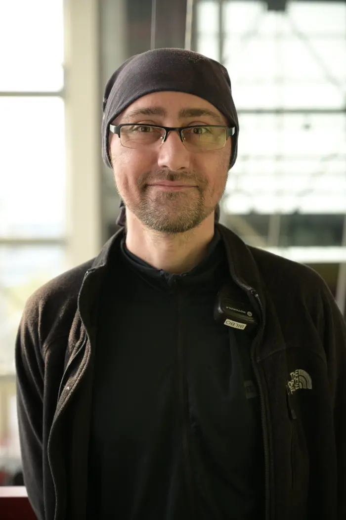 Un homme portant des lunettes, un foulard sombre et une veste North Face noire sourit à l'appareil photo à l'intérieur ; des fenêtres floues et de la lumière en arrière-plan capturent l'humeur détendue de Bilbo Rochette.