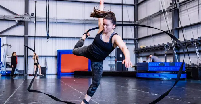 Une femme exécute un mouvement de danse dynamique avec un long ruban noir dans un gymnase d'un centre de recherche, en équilibre sur une jambe tout en étendant ses bras et ses jambes, entourée d'équipements de gymnastique et d'autres personnes à l'arrière-plan.