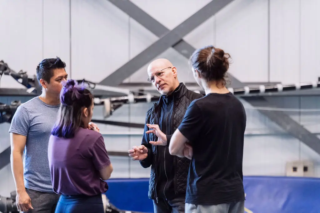 Quatre diplômés en enseignement de l'ENC se tiennent en groupe à l'intérieur, engagés dans une conversation. Une personne vêtue d'une veste noire fait un geste tandis que les autres écoutent attentivement. Des équipements industriels et des poutres métalliques sont visibles à l'arrière-plan.