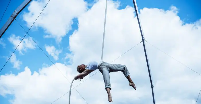 Une personne vêtue d'un pantalon gris et d'une chemise blanche effectue un numéro d'acrobatie aérienne sur une corde suspendue, dans un ciel bleu vif parsemé de nuages, incarnant l'esprit de mardi je donne à l'ÉNC.