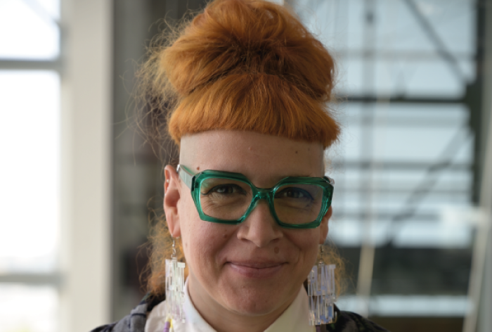 Anna-Karyna Barlati, cheveux orange vif coiffés en chignon haut et côtés rasés, porte de grosses lunettes vertes, des boucles d'oreilles et une chemise blanche, souriant légèrement dans un décor intérieur moderne.