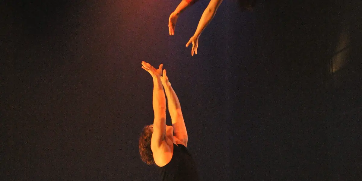 Un acrobate masculin, Antoine Auger, sur scène, les bras levés, attrape Geneviève Morin en plein vol, tous deux éclairés par des projecteurs colorés, avec un canapé blanc en arrière-plan.