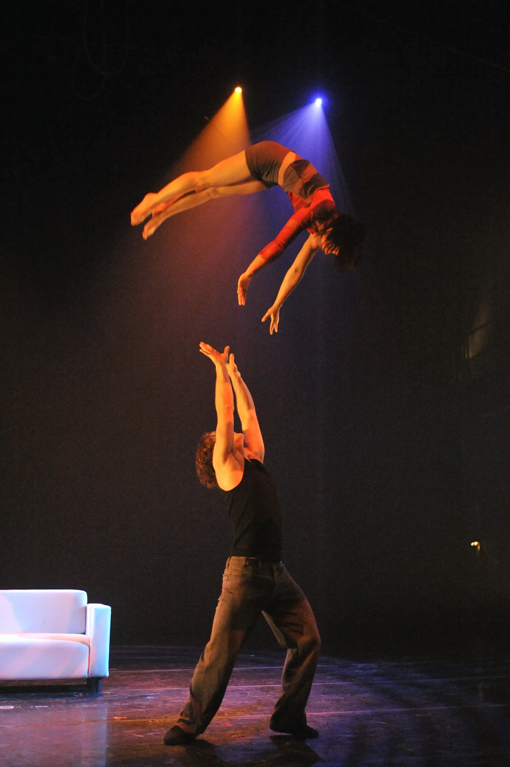 Un acrobate masculin, Antoine Auger, sur scène, les bras levés, attrape Geneviève Morin en plein vol, tous deux éclairés par des projecteurs colorés, avec un canapé blanc en arrière-plan.
