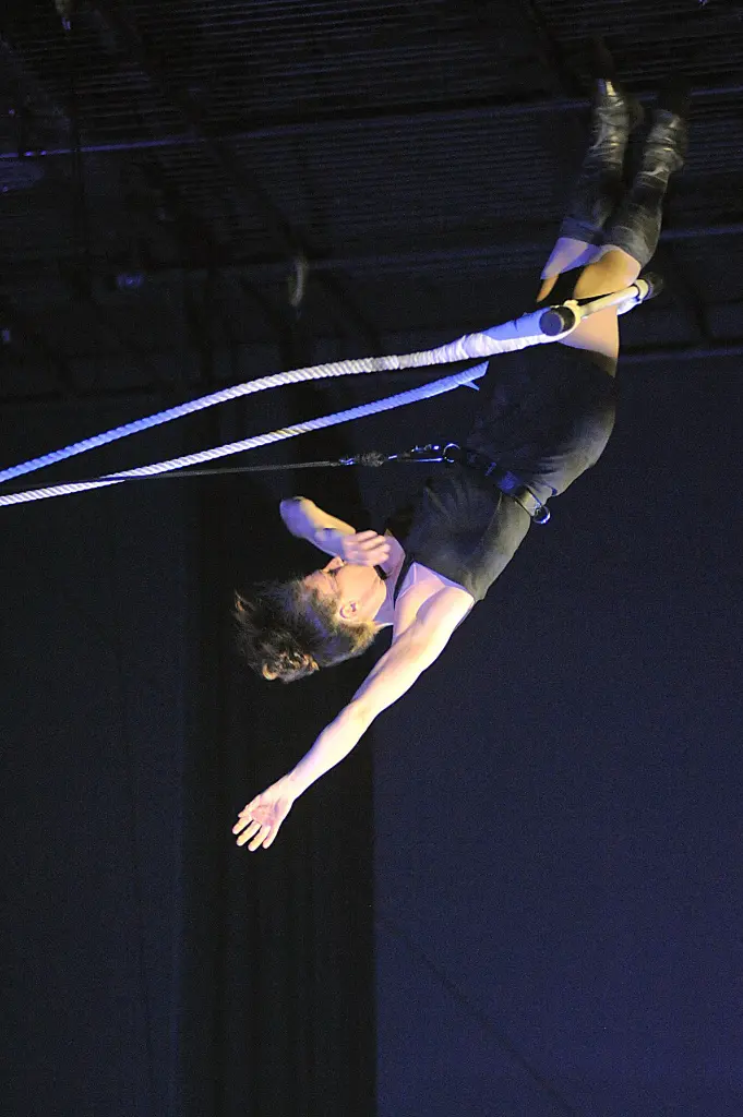 Aurélie Fléchais, vêtue d'une tenue noire et de bottes, exécute un numéro de trapèze aérien, suspendue à l'envers dans les airs, un bras tendu et l'autre plié, sur un fond sombre.