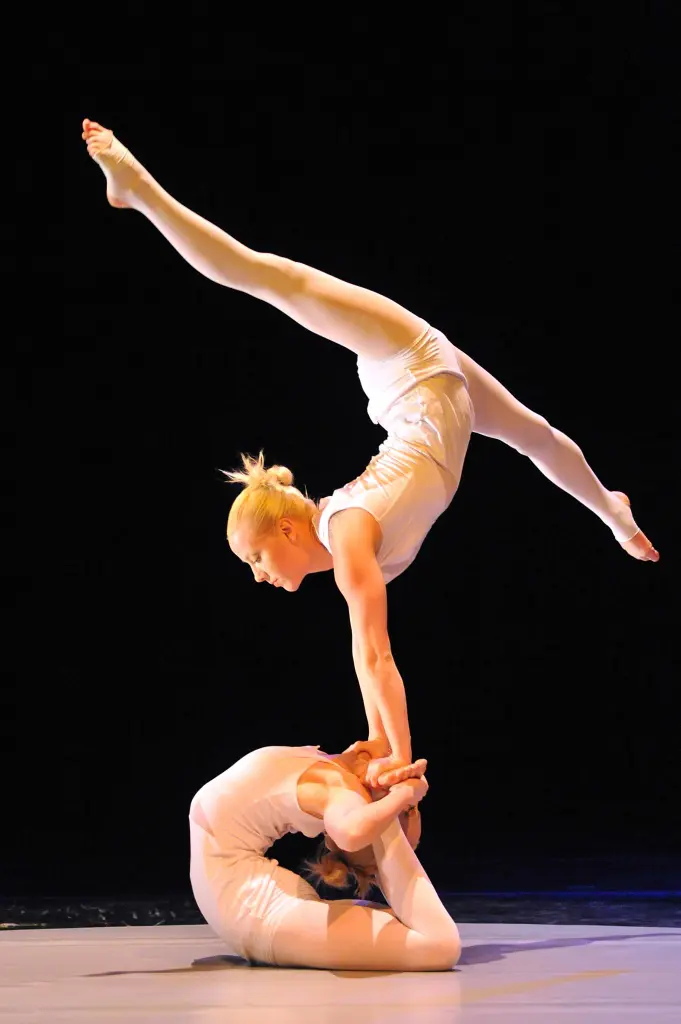 Jenny et Sara Haglund, vêtues de costumes blancs, réalisent un étonnant numéro d'équilibre. L'une des acrobates est allongée sur le sol en se tenant les pieds, tandis que l'autre se tient en équilibre sur le dos dans une fente. Un arrière-plan sombre met en valeur leur impressionnante pose.
