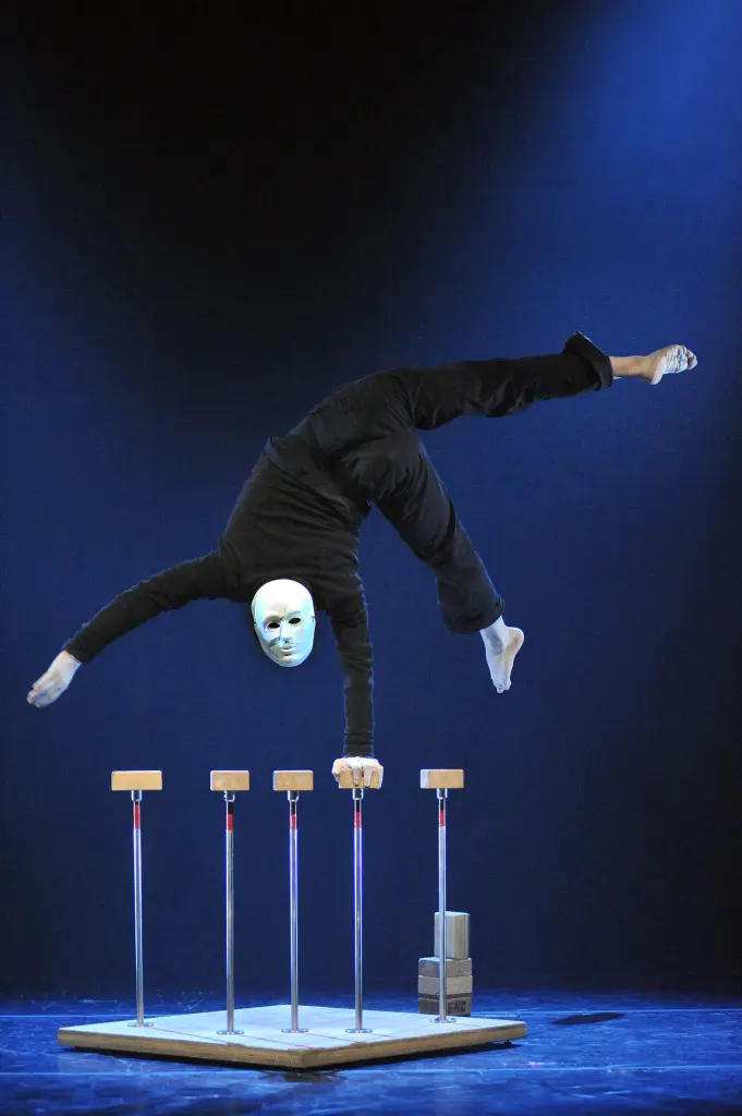 Un interprète vêtu de noir et portant un masque blanc se tient en équilibre sur une main au sommet de cannes, étendant ses jambes en l'air sous un éclairage de scène bleu spectaculaire - une démonstration impressionnante de Naël Jammal.