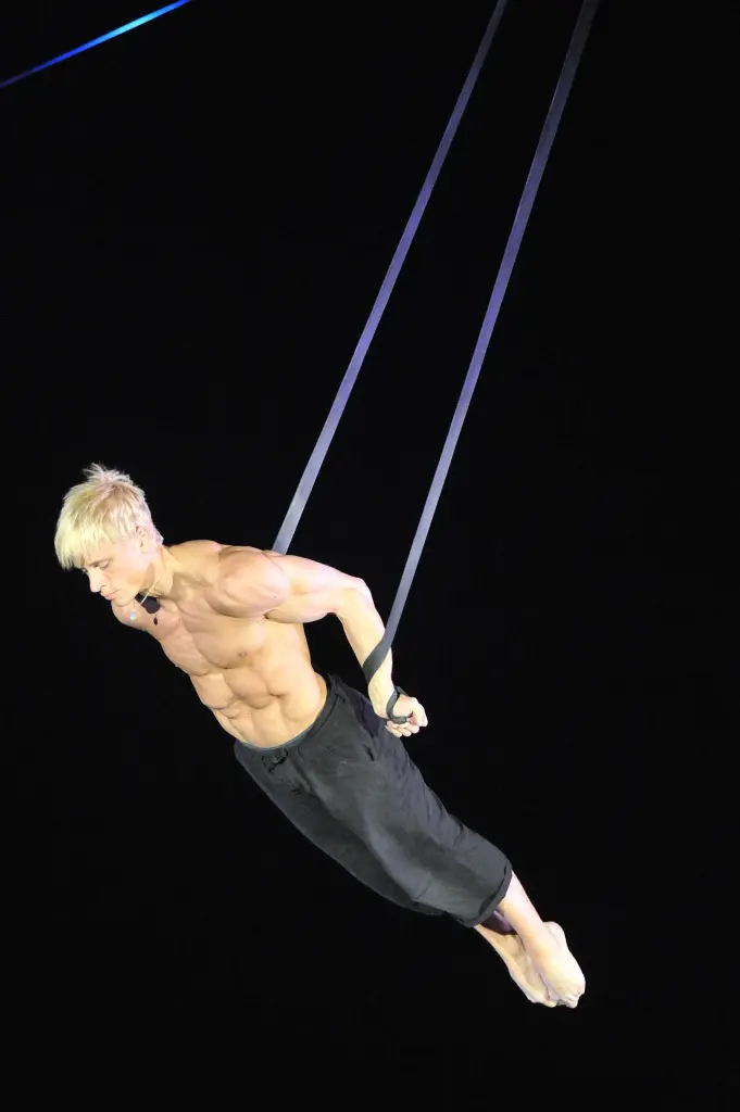Un homme musclé aux cheveux blonds, identifié comme Hampus Jansson, exécute une routine aérienne sur des anneaux de gymnastique, en tenant son corps à l'horizontale, les bras tendus, sur un fond noir. Il porte un pantalon noir et semble concentré.