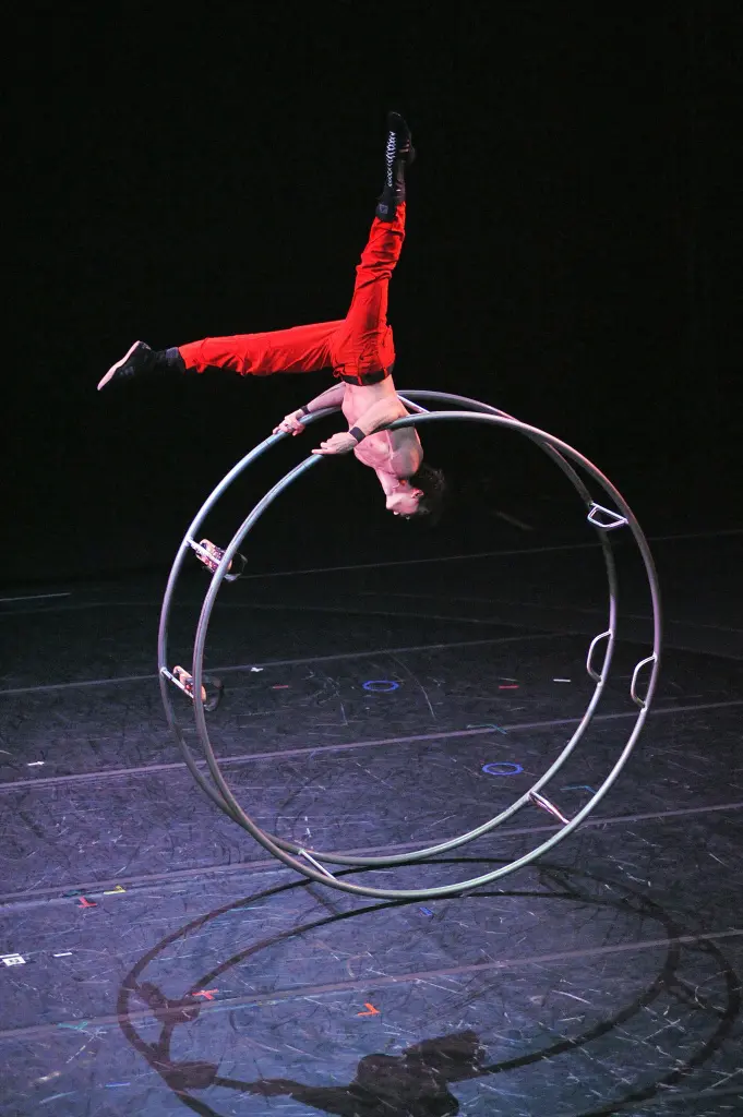 Un artiste vêtu d'un pantalon rouge et d'un haut de forme, rappelant le style audacieux de Frédéric Lemieux-Cormier, se tient en équilibre à l'envers sur une grande roue métallique, une jambe tendue vers le haut, au cours d'un numéro d'acrobatie sur une scène obscure.