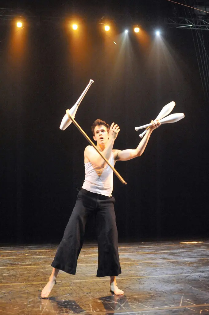 Un artiste pieds nus, vêtu d'un débardeur blanc et d'un pantalon noir, rappelant Florent Lestage, jongle avec des massues sur scène, sous des projecteurs lumineux, en se concentrant sur l'une d'entre elles, posée en équilibre sur un bâton.