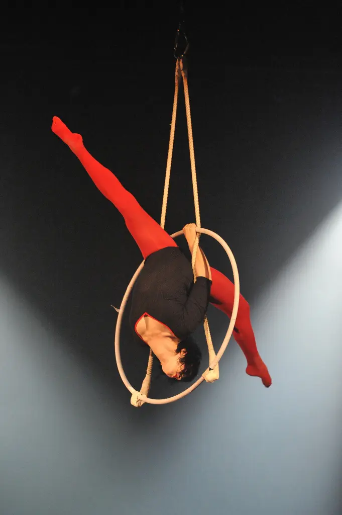 Maria Terentieva, en collants rouges et haut noir, prend une pose acrobatique sur un cerceau suspendu, étendant une jambe en hauteur tout en saisissant le cerceau avec ses mains, sur un fond sombre et éclairé par des projecteurs.