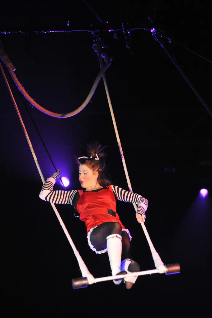 Une artiste de cirque en costume rouge et manches rayées - rappelant Anne-Fay Audet Johnston - se balance sur un trapèze sous les lumières de la scène, avec un arrière-plan sombre et des cordes suspendues au-dessus.