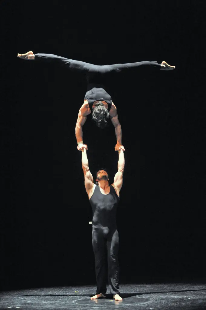 Gregory Morgan Arsenal Romagosa et Yannick Thomas se produisent sur scène ; l'un se tient debout en tenant l'autre, qui est à l'envers et en équilibre sur les mains, les jambes écartées. Tous deux portent des tenues noires sur un fond sombre.