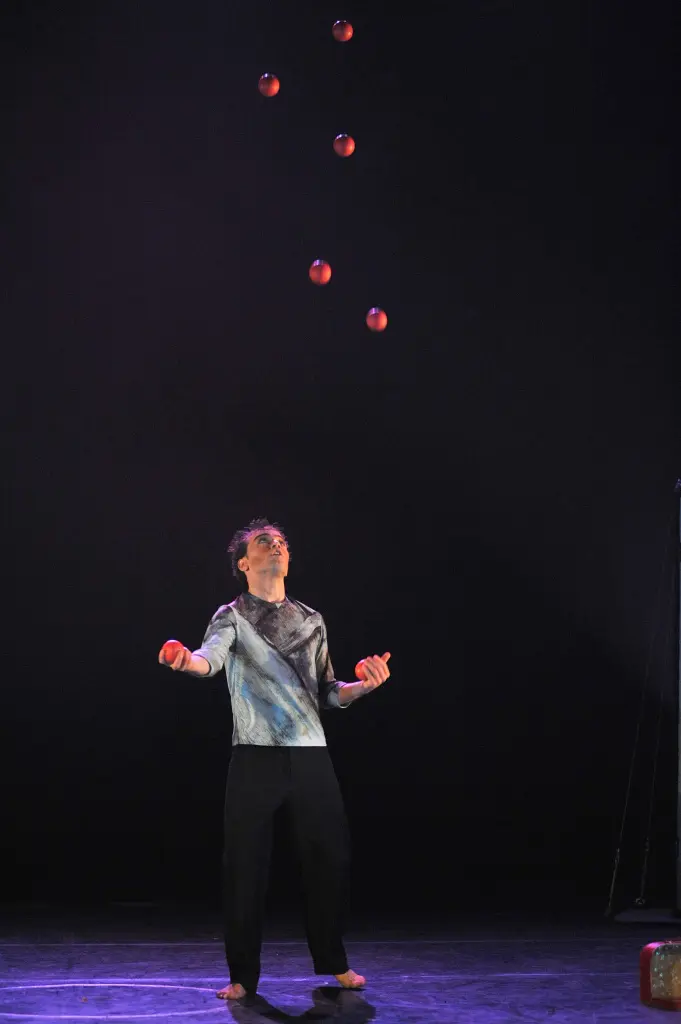 Un artiste aux pieds nus, Giulio Lanzafame, se tient sous l'éclairage de la scène et jongle avec sept balles rouges en l'air, regardant attentivement sur un fond sombre.