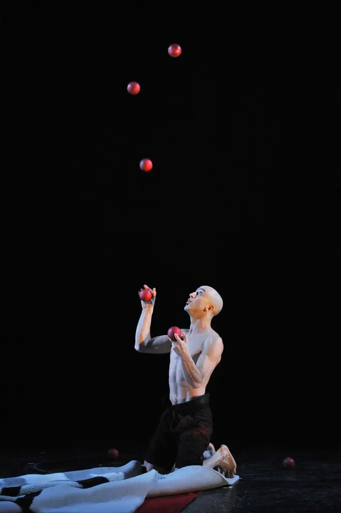 David Menes Rodriguez, torse nu, est agenouillé sur scène, jonglant avec expertise avec cinq balles rouges dans les airs sur un fond sombre, quelques balles reposant sur le sol à proximité.