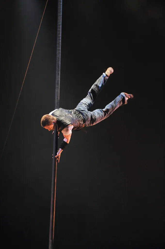 Fletcher Sanchez, vêtu de vêtements sombres, fait un numéro d'équilibriste à une main sur un poteau vertical, étendant son corps à l'horizontale avec un bras agrippé au poteau, le tout sur un fond sombre dramatique.