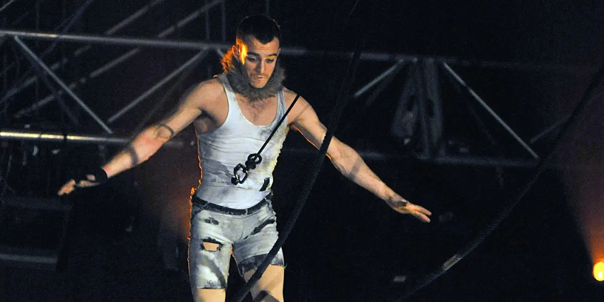 Anthony Weiss, un acrobate masculin vêtu d'un costume argenté, se tient en équilibre sur une corde raide, les bras tendus pour garder l'équilibre, l'éclairage de la scène et le gréement étant visibles à l'arrière-plan.