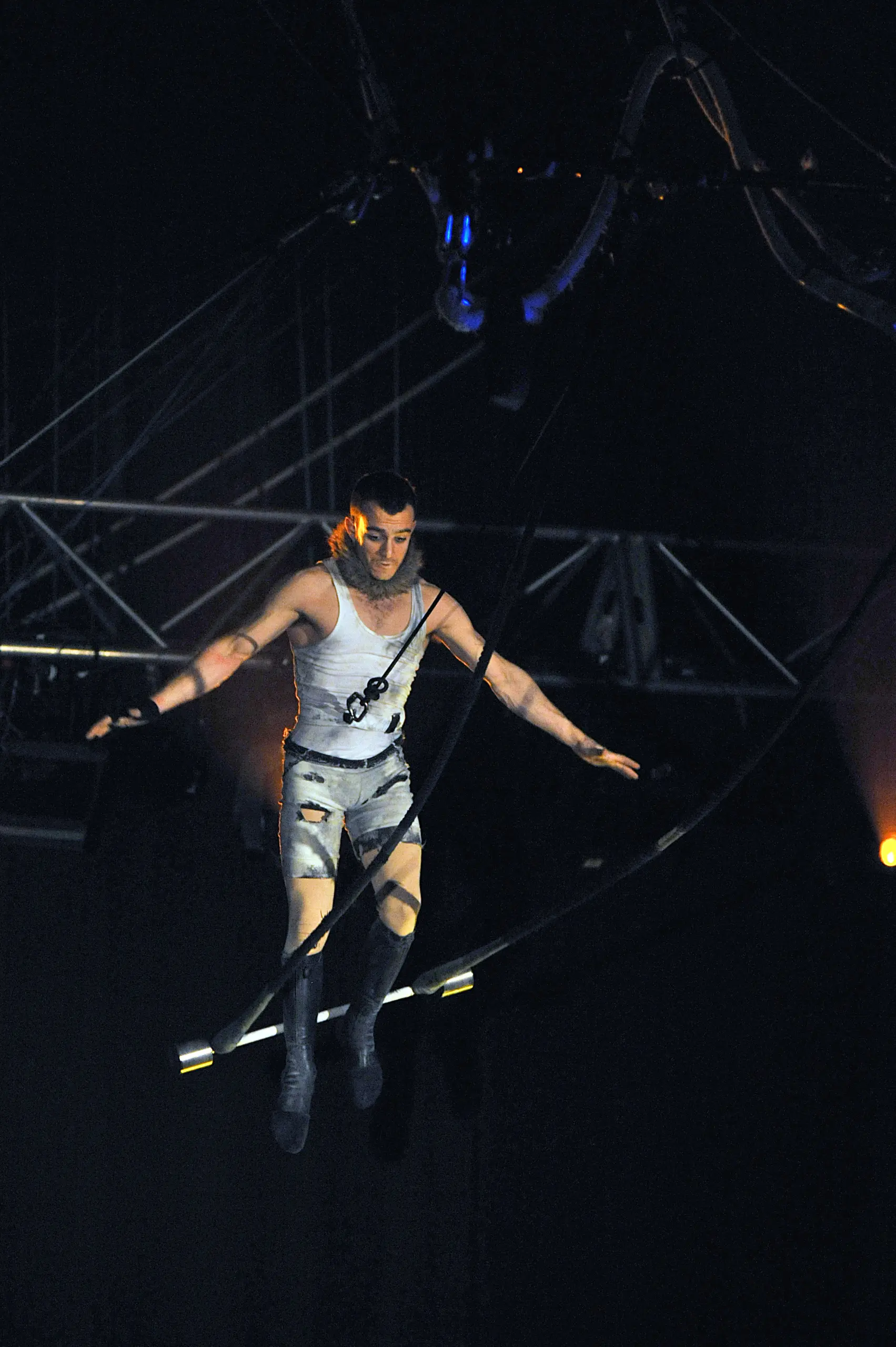 Anthony Weiss, un acrobate masculin vêtu d'un costume argenté, se tient en équilibre sur une corde raide, les bras tendus pour garder l'équilibre, l'éclairage de la scène et le gréement étant visibles à l'arrière-plan.