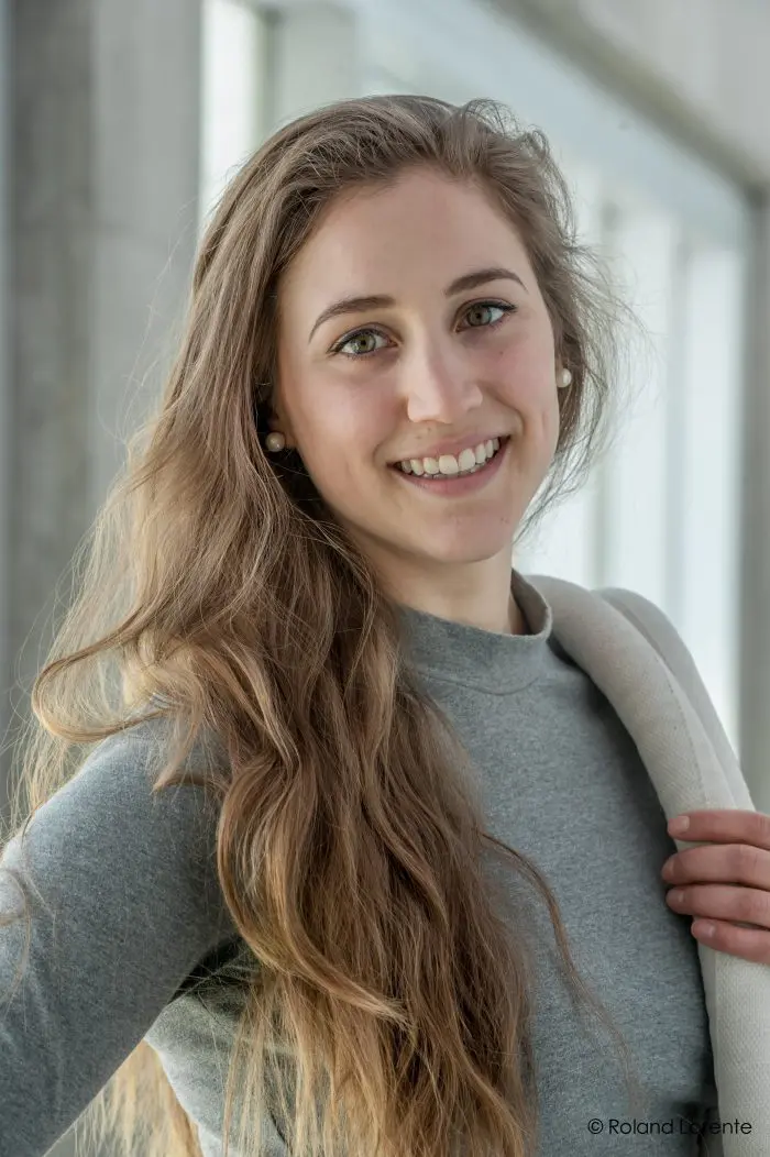 Selene Ballesteros-Minguer, une jeune femme aux longs cheveux bruns ondulés, sourit en portant un pull gris et en tenant une lanière blanche sur son épaule. Elle se tient à l'intérieur sur un fond moderne flou.