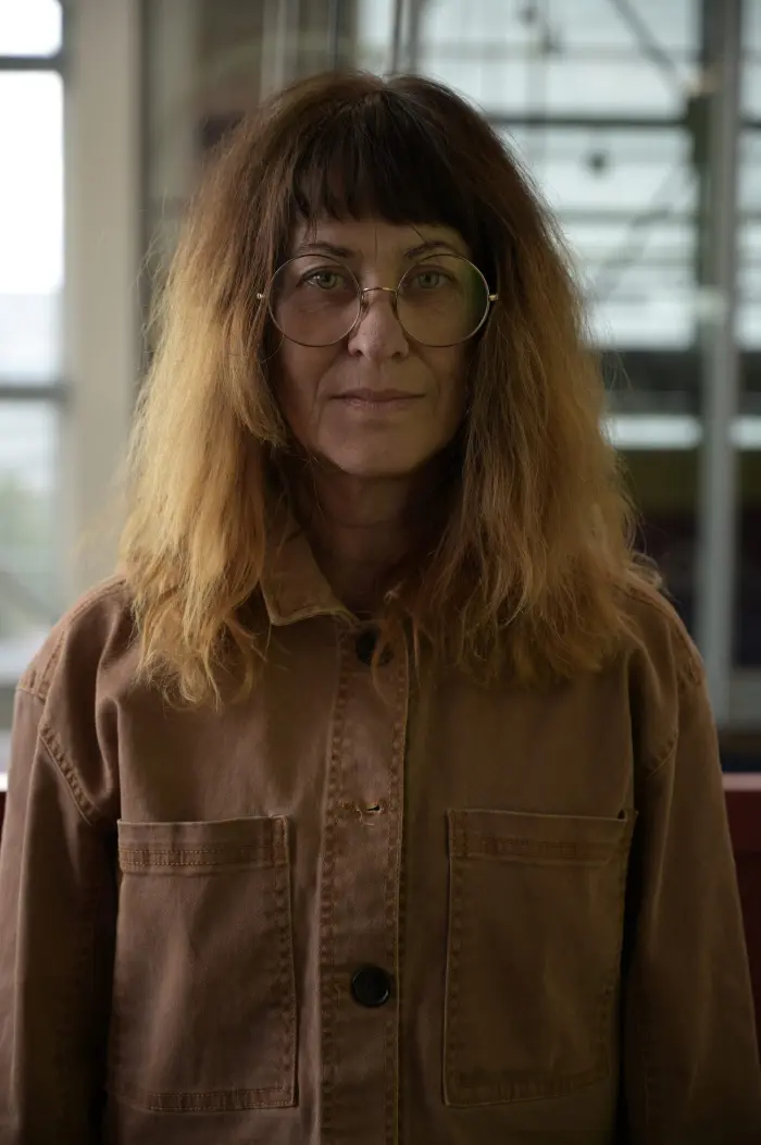 Une femme aux longs cheveux bruns ondulés et aux lunettes rondes se tient à l'intérieur, vêtue d'une veste boutonnée marron. Elle regarde directement l'appareil photo avec une expression neutre. L'arrière-plan légèrement flou complète ce portrait inspiré de Johanne Madore.
