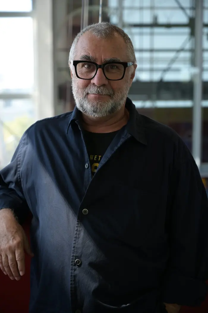 Un homme d'âge moyen aux cheveux gris, à la barbe et aux lunettes noires se tient à l'intérieur - il s'agit de Rénald Laurin - dans une chemise boutonnée et un t-shirt sombres, posant avec une main sur une surface, sur fond de grandes fenêtres.