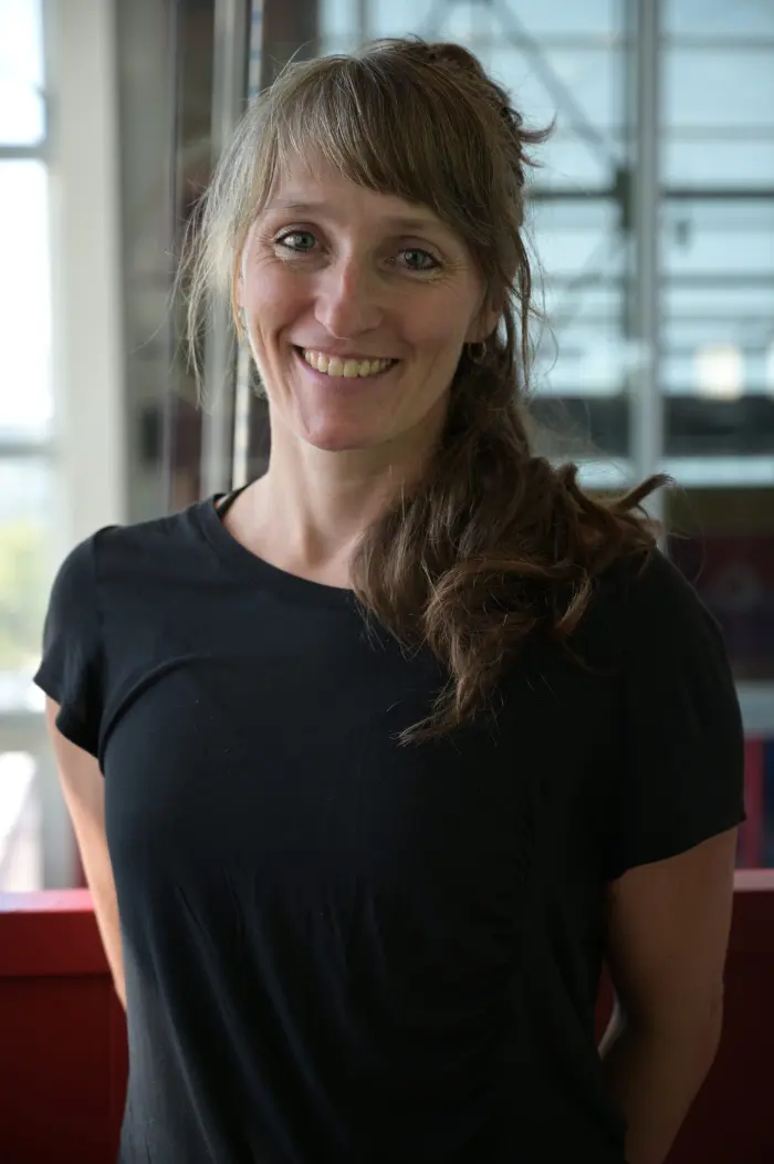 Véronique Thibeault, une femme aux longs cheveux châtain clair attachés sur le côté et vêtue d'un t-shirt noir, sourit chaleureusement alors qu'elle se tient à l'intérieur, avec des fenêtres en verre et une balustrade rouge en arrière-plan.