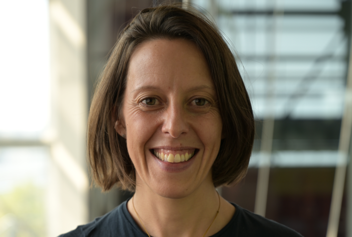 Vanessa Audebert, une femme aux cheveux bruns courts, vêtue d'une chemise sombre et d'un collier en or, sourit à l'appareil photo dans un intérieur très éclairé, avec de grandes fenêtres en arrière-plan.