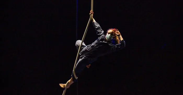 Une personne portant un masque et des vêtements sombres exécute un numéro aérien, s'agrippant à une corde verticale en plein vol sur un fond sombre - reprenant l'esprit d'audace vu dans la série documentaire n'arrête pas ton cirque !.