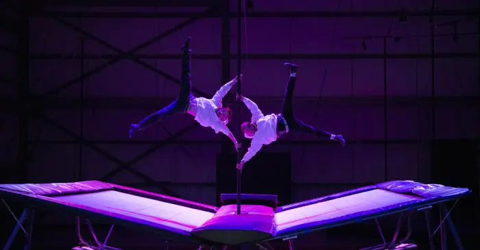 Deux acrobates en chemise blanche réalisent un équilibre synchronisé sur un poteau, en équilibre au-dessus de trampolines, sous un éclairage violet spectaculaire dans une arène intérieure lors de l'événement "Subvention pendant 5 ans pour le CRITAC".