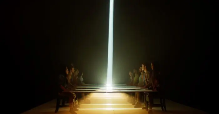 Dans une pièce faiblement éclairée, une rangée de personnes est assise de part et d'autre d'une longue table, face à face. Un faisceau lumineux vertical brille au centre, illuminant la scène de façon spectaculaire - en écho à l'atmosphère de la somme de nos rêves, le film des finissant-e-s 2021.