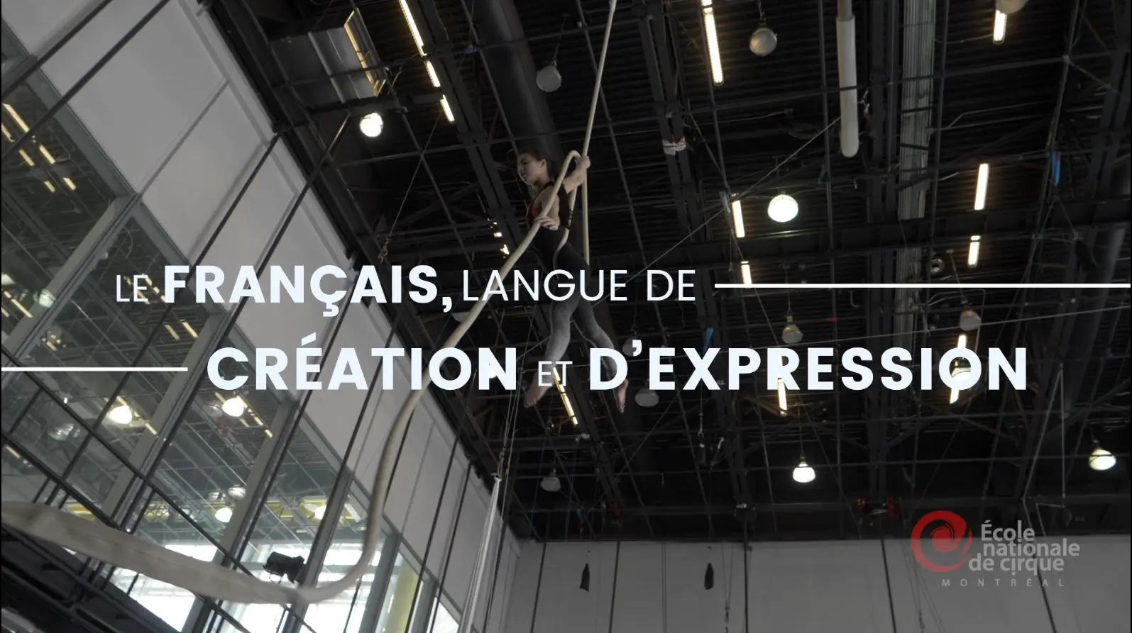 Artiste aérien évoluant en hauteur sur des cordes dans un grand gymnase, lumières brillantes. Le texte en français superposé est le suivant : "Le français, langue de création et d'expression" : Le français, langue de création et d'expression, soulignant la campagne commune de promotion du français. Logo de l'École nationale de cirque Montréal dans le coin.