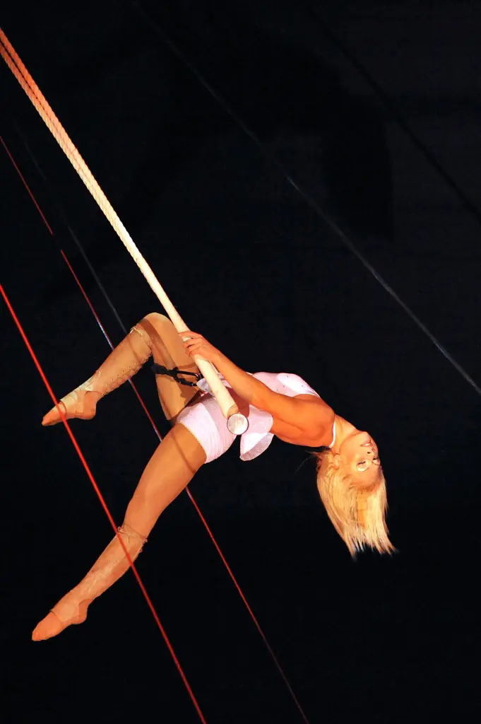 Une acrobate, Uuve-hanna Jansson, aux cheveux blonds, exécute un numéro aérien, suspendue la tête en bas à une corde épaisse sur un fond sombre, vêtue d'un costume et de collants de couleur claire.