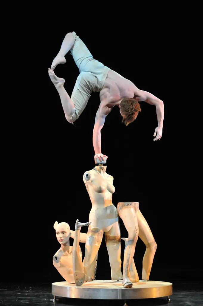 Un acrobate masculin, rappelant une composition de Philip Rosenberg, se tient en équilibre sur une main, la tête en bas, au-dessus d'un groupe de mannequins sans tête, vêtus d'un pantalon de couleur claire, sur un fond noir et un éclairage de scène dramatique.