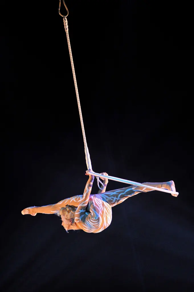 L'artiste aérienne Sabrina Aganier est suspendue à une corde et exécute une pose acrobatique en plein vol. Elle arque le dos et s'accroche à la corde avec les deux mains sur un fond sombre.