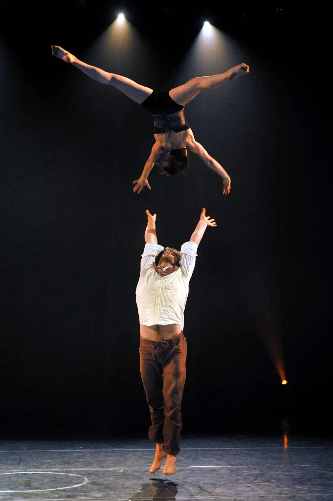 Un acrobate pieds nus fait tenir un autre acrobate en équilibre sur ses mains levées. Valérie Benoit-Charbonneau et Mason Ames exécutent un split sous un éclairage de scène dramatique, mis en valeur sur un fond sombre.