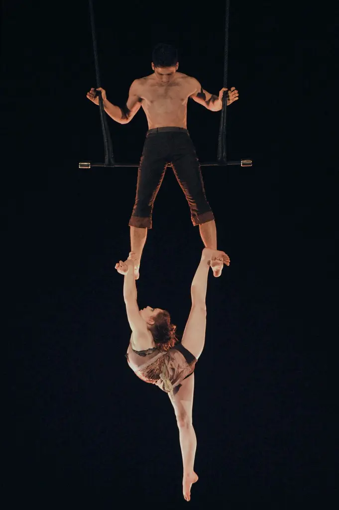 Un acrobate, Louis-David Simoneau, se tient sur un trapèze, tenant la main de Rosalie Ducharme, qui est suspendue à l'envers sous lui dans un impressionnant dédoublement sur un fond sombre.