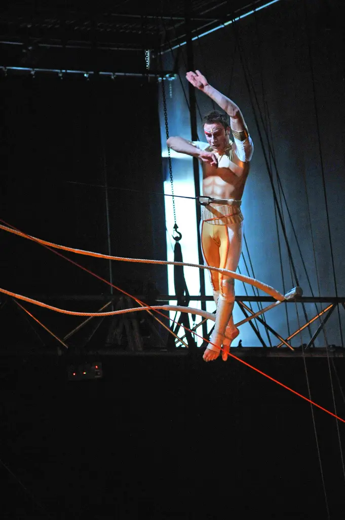 Un interprète, qui rappelle Noé Robert, vêtu d'un costume clair, se tient en équilibre sur une corde raide au-dessus de la scène, les bras levés au milieu de cordes et de gréements. L'arrière-plan sombre et dramatique renforce le suspense de ce numéro à couper le souffle.