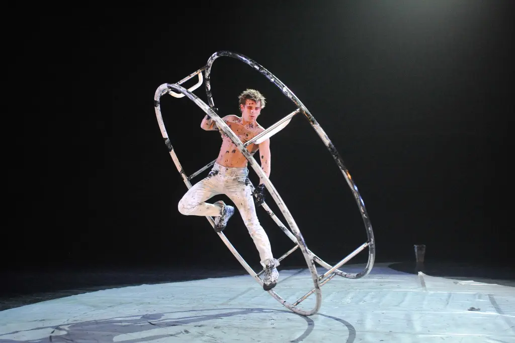 Un artiste masculin, Julien Silliau, se tient habilement en équilibre dans une grande roue Cyr en métal sur une scène faiblement éclairée, vêtu d'un pantalon blanc et d'un torse nu peint, un projecteur mettant en valeur sa pose dynamique.