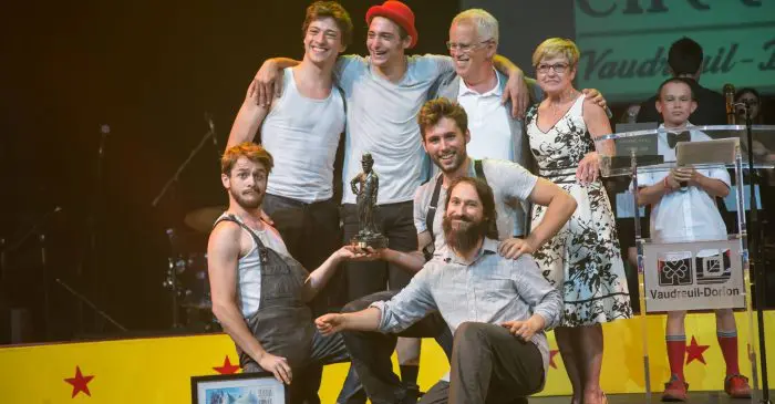 Un groupe d'artistes pose sur scène, souriant, certains agenouillés et d'autres debout, tenant une photo encadrée et un trophée, Machine de Cirque remporte le Trophée Charlie-Chaplin, avec deux adultes plus âgés et un enfant à côté d'un podium à l'arrière-plan.
