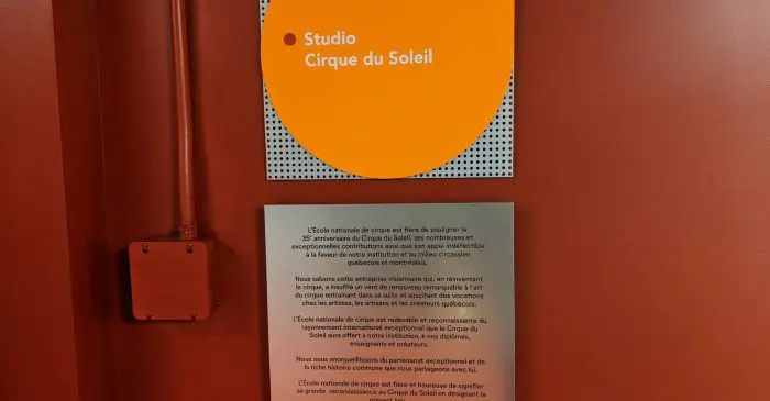 Sur un mur orange, un panneau circulaire orange indique " Studio de l'ÉNC nommé le " Studio Cirque du Soleil " au-dessus d'une plaque blanche avec un texte en français. Une boîte de service orange est installée à gauche sous l'enseigne.