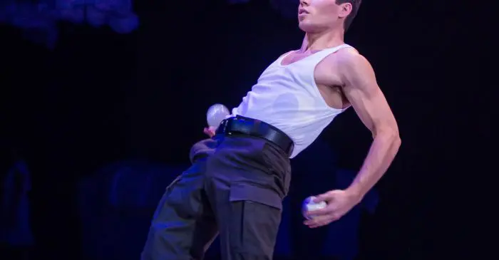 Un homme pieds nus, vêtu d'un débardeur blanc et d'un pantalon sombre, jongle avec cinq balles blanches sur scène, en se penchant en arrière avec concentration. La scène fait écho à l'habileté de plusieurs diplômé-e-s de l'ECN ont reçu des prix, sur un fond sombre à l'éclairage tamisé.