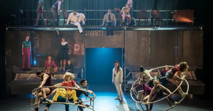 Un groupe d'artistes sur scène, certains debout ou assis sur une plate-forme surélevée et d'autres interagissant avec de grandes structures métalliques circulaires, sous un éclairage dramatique dans un cadre théâtral, alors que sept créateurs prendront part à la semaine de création 2020 de l'École nationale de cirque.