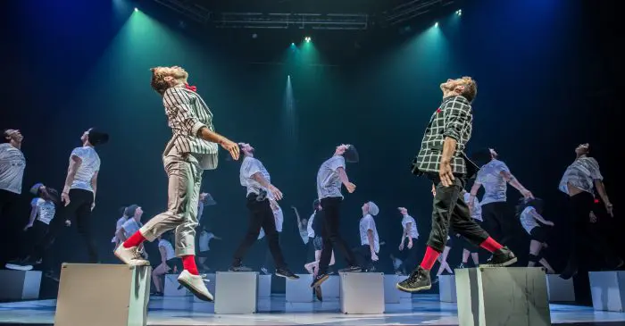 Un groupe de danseurs en tenue coordonnée bondit sur scène, éclairé par des projecteurs bleus et violets. À l'avant, mis en valeur par des cubes, se trouvent deux danseurs - des vedettes des spectacles annuels 2016 de metteur-euse-s en piste.