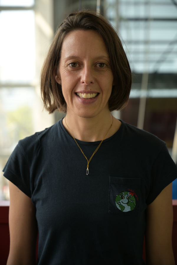 Une femme aux cheveux bruns courts, vêtue d'un t-shirt foncé avec un petit écusson de la Terre sur la poche et un collier à pendentif, se tient à l'intérieur, souriant à l'appareil photo sur fond de fenêtre floue - il pourrait s'agir de Vanessa Audebert.