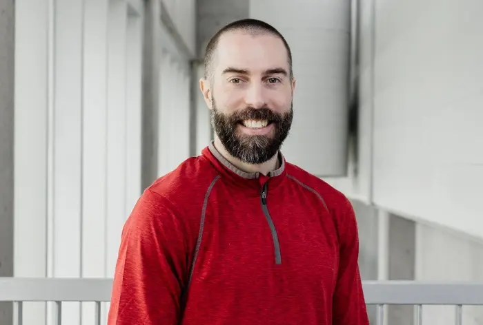 Un homme barbu, Chris Gatti, vêtu d'un pull-over rouge, se tient à l'intérieur et sourit à la caméra. L'arrière-plan est constitué de murs blancs et de fenêtres baignées de lumière naturelle.