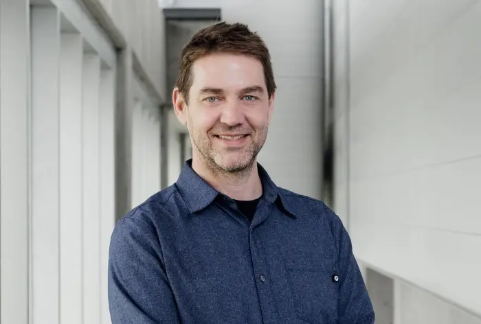 Emmanuel Bochud, un homme aux cheveux bruns courts et à la barbe, sourit en croisant les bras, vêtu d'une chemise bleue boutonnée, dans un couloir moderne et lumineux aux murs clairs.