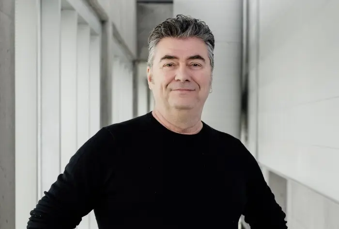 Fred Gérard, un homme d'âge moyen aux cheveux gris, se tient à l'intérieur contre un mur blanc, vêtu d'une chemise noire à manches longues, souriant à l'appareil photo avec assurance, les mains sur les hanches.