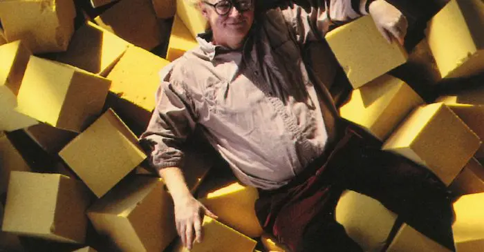Un homme à lunettes et aux cheveux bouclés est allongé, souriant et détendu sur une grande pile de cubes de mousse jaune. Vêtu d'une chemise rose pâle et d'un pantalon sombre, il dégage le charme décontracté d'un portrait de Jan Rok Archard.