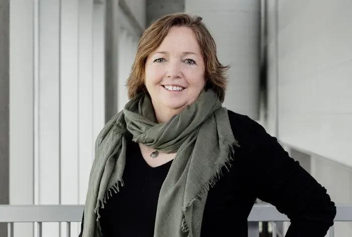Line Giasson, une femme aux cheveux bruns longs comme le menton, sourit à l'appareil photo. Elle porte un haut sombre et une grande écharpe vert olive. Elle se tient à l'intérieur sur un fond clair et moderne.