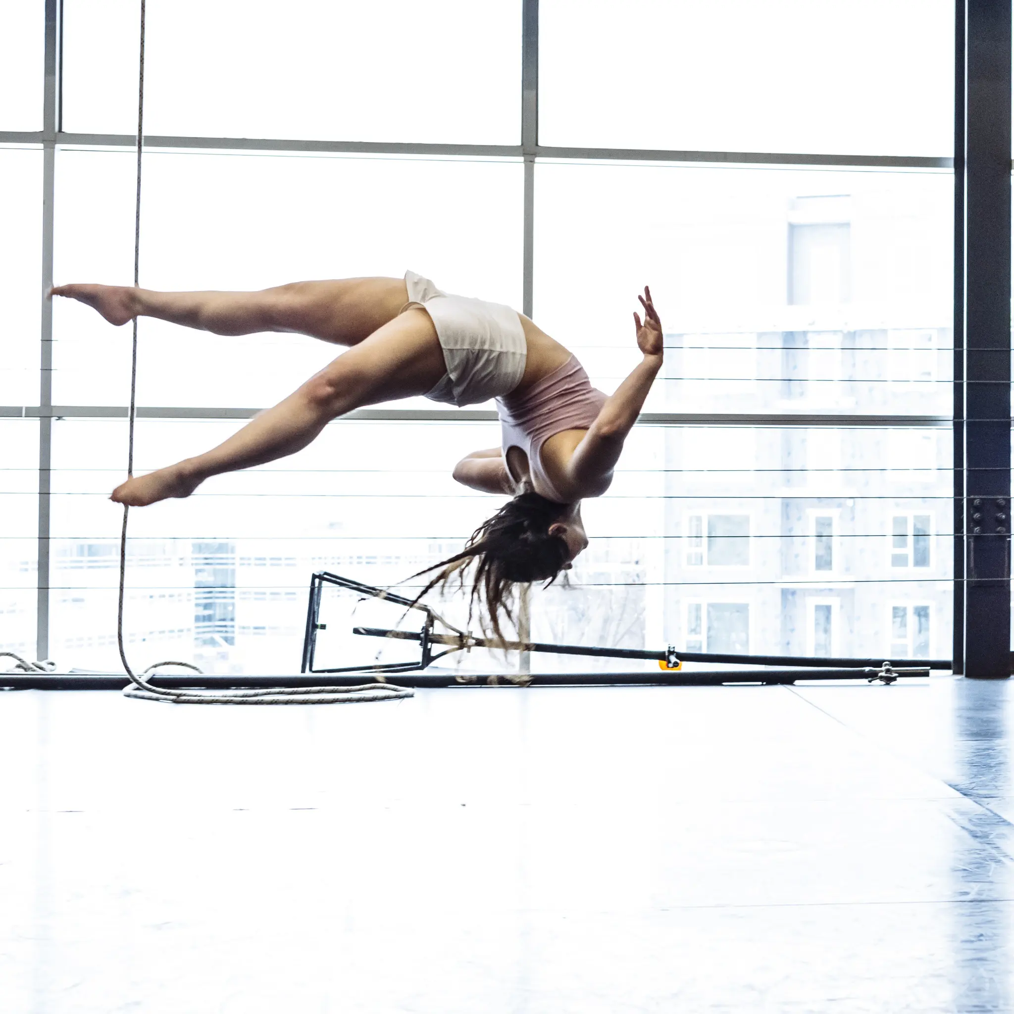 Une gymnaste en plein vol exécute un saut périlleux arrière dans un espace intérieur lumineux au Concours Élève d'un jour, où l'ENC accueille des 9 à 17 ans. Elle porte un haut rose pâle et un short blanc, son corps s'étendant à l'horizontale pendant qu'elle tourne.
