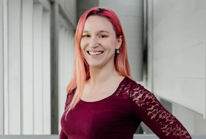 Marion Cossin, aux longs cheveux roses et raides, sourit dans un couloir lumineux. Elle porte un haut bordeaux avec des manches en dentelle et a une main sur la hanche. Des murs blancs et de grandes fenêtres forment l'arrière-plan.